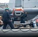 The image shows U.S. Navy personnel conducting deck operations aboard a naval vessel, likely during a boat handling or equipment maintenance evolution. Several sailors wearing different colored hard hats are working together on what appears to be a rigid hull inflatable boat (RHIB) or similar small craft secured to the ship's side. An American flag is visible in the background, confirming the U.S. military context.
