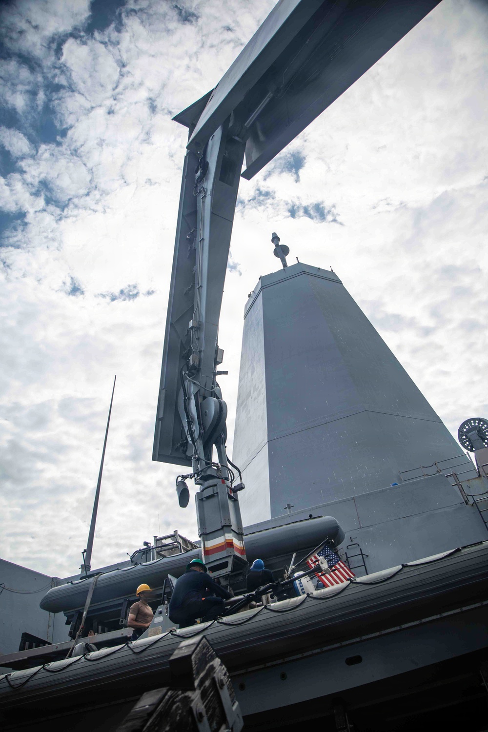 A low-angle upward shot taken aboard what appears to be a U.S. Navy warship, likely a Zumwalt-class destroyer, showing the distinctive angular superstructure and a crane or davit arm extending overhead. Several crew members wearing hard hats are visible on deck near what appears to be a rigid inflatable boat (RIB). An American flag is visible on the vessel's side.