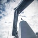A low-angle upward shot taken aboard what appears to be a U.S. Navy warship, likely a Zumwalt-class destroyer, showing the distinctive angular superstructure and a crane or davit arm extending overhead. Several crew members wearing hard hats are visible on deck near what appears to be a rigid inflatable boat (RIB). An American flag is visible on the vessel's side.