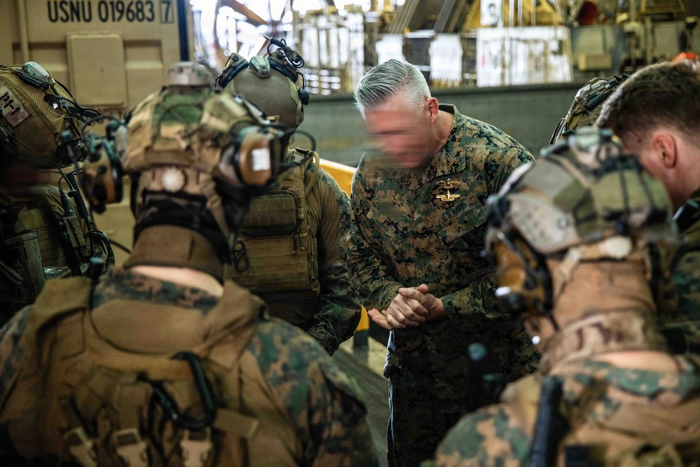 A group of military personnel in MARPAT camouflage uniforms are gathered in what appears to be the interior of a naval vessel or military transport. A senior officer with gray hair, wearing a badge on his chest, appears to be in a huddle or briefing with fully equipped combat-ready personnel. The personnel are wearing full tactical gear including helmets, body armor, and communication equipment.