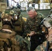 A group of military personnel in MARPAT camouflage uniforms are gathered in what appears to be the interior of a naval vessel or military transport. A senior officer with gray hair, wearing a badge on his chest, appears to be in a huddle or briefing with fully equipped combat-ready personnel. The personnel are wearing full tactical gear including helmets, body armor, and communication equipment.