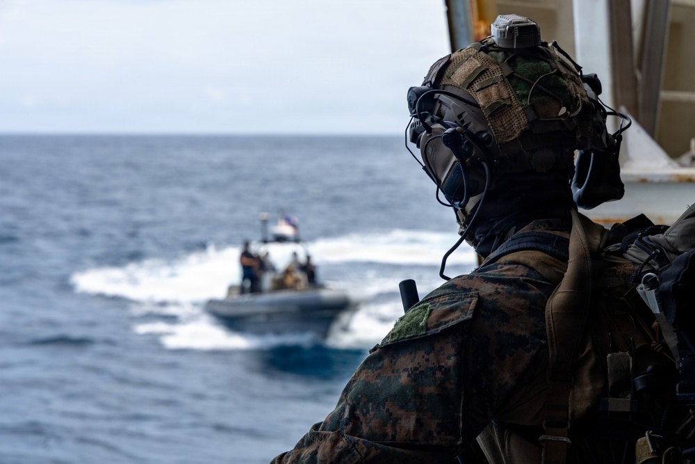A heavily equipped military personnel in MARPAT (Marine pattern) camouflage uniform observes a rigid-hull inflatable boat (RHIB) operating on open ocean waters. The soldier is wearing a tactical helmet with communications equipment and is positioned on what appears to be the deck or ramp of a larger vessel. The RHIB in the background carries several personnel and is flying a small flag.