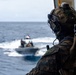 A heavily equipped military personnel in MARPAT (Marine pattern) camouflage uniform observes a rigid-hull inflatable boat (RHIB) operating on open ocean waters. The soldier is wearing a tactical helmet with communications equipment and is positioned on what appears to be the deck or ramp of a larger vessel. The RHIB in the background carries several personnel and is flying a small flag.