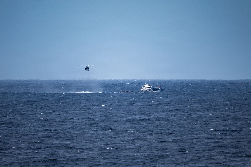 A maritime scene showing a helicopter hovering low over open ocean near what appears to be a smaller dark vessel, with a larger white motor yacht visible nearby. Smoke or spray is visible near the water surface beneath the helicopter, suggesting an active operation. The scene appears to depict a maritime interdiction, rescue, or law enforcement operation at sea.