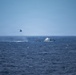 A maritime scene showing a helicopter hovering low over open ocean near what appears to be a smaller dark vessel, with a larger white motor yacht visible nearby. Smoke or spray is visible near the water surface beneath the helicopter, suggesting an active operation. The scene appears to depict a maritime interdiction, rescue, or law enforcement operation at sea.