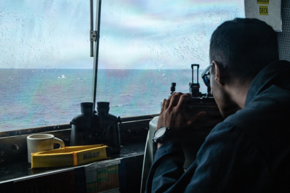 A person is seated inside what appears to be a ship's bridge or observation deck, looking through an optical sighting device toward the open sea. The rain-streaked window and overcast sky suggest wet weather conditions at sea. Navigation and observation equipment is visible on the console in front of them.