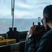 A person is seated inside what appears to be a ship's bridge or observation deck, looking through an optical sighting device toward the open sea. The rain-streaked window and overcast sky suggest wet weather conditions at sea. Navigation and observation equipment is visible on the console in front of them.