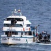 A law enforcement or military interdiction operation is underway at sea, with a rigid inflatable boat (RIB) carrying armed personnel alongside a larger white vessel named 'Manta'. The larger vessel displays what appears to be an Ecuadorian flag and registration markings. Personnel in tactical gear are visible transferring or boarding between the two vessels.