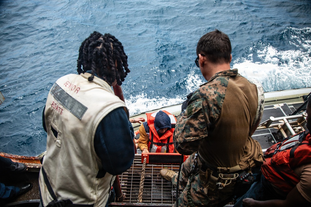 The image shows a maritime rescue or boarding operation taking place on the deck of a naval vessel. A person wearing an orange life vest is being assisted up a ladder or gangway from the water level by military and naval personnel. Two individuals are prominently visible in the foreground — one wearing a 'Senior Boats' vest and another in U.S. Marine Corps MARPAT camouflage uniform.