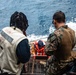 The image shows a maritime rescue or boarding operation taking place on the deck of a naval vessel. A person wearing an orange life vest is being assisted up a ladder or gangway from the water level by military and naval personnel. Two individuals are prominently visible in the foreground — one wearing a 'Senior Boats' vest and another in U.S. Marine Corps MARPAT camouflage uniform.