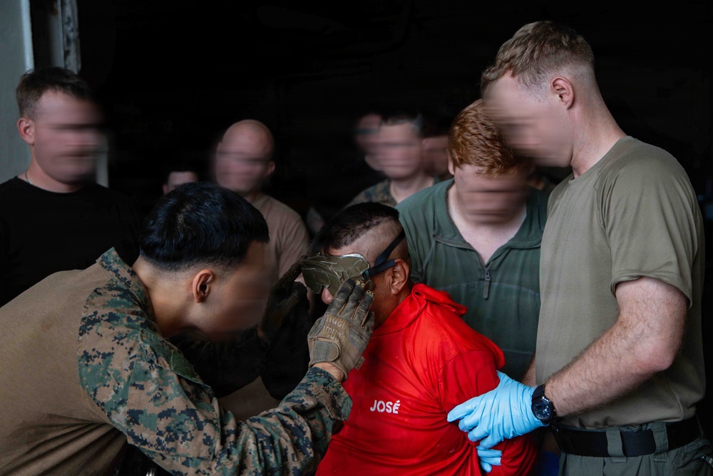 A military training or operational scenario showing personnel in uniform conducting a detainee handling or search procedure. A service member in camouflage uniform and tactical gloves is examining or restraining an individual wearing a red shirt labeled 'JOSÉ'. Multiple other personnel in military/civilian attire observe or assist in the background.