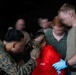 A military training or operational scenario showing personnel in uniform conducting a detainee handling or search procedure. A service member in camouflage uniform and tactical gloves is examining or restraining an individual wearing a red shirt labeled 'JOSÉ'. Multiple other personnel in military/civilian attire observe or assist in the background.