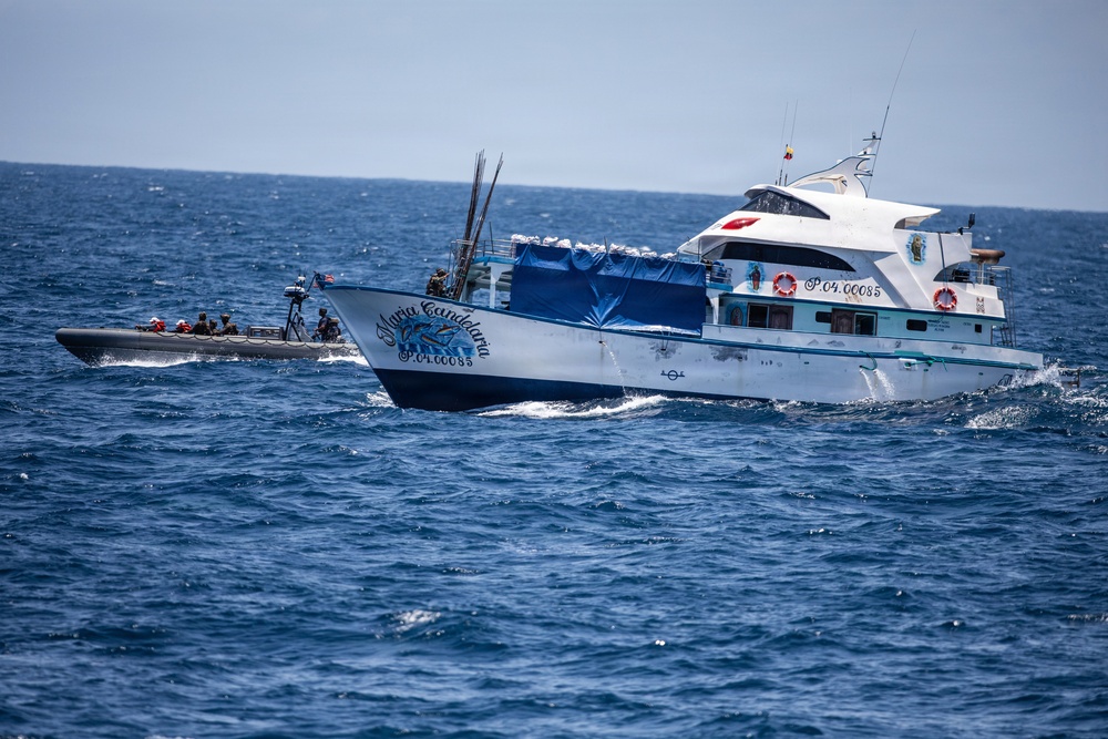 A white and blue vessel named 'Maria Candelaria' with registration number P.04.00085 is underway on open ocean waters. A smaller rigid inflatable boat (RIB) with several personnel aboard is positioned alongside the larger vessel, suggesting an interdiction or boarding operation. A flag is visible on the RIB, and a small flag is also present on the larger vessel's superstructure.