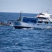 A white and blue vessel named 'Maria Candelaria' with registration number P.04.00085 is underway on open ocean waters. A smaller rigid inflatable boat (RIB) with several personnel aboard is positioned alongside the larger vessel, suggesting an interdiction or boarding operation. A flag is visible on the RIB, and a small flag is also present on the larger vessel's superstructure.