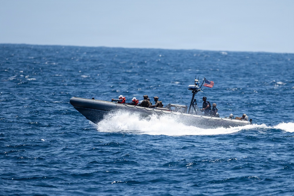 A rigid-hull inflatable boat (RHIB) is underway at high speed on open blue ocean water. The vessel carries multiple personnel in military/tactical gear and displays a United States flag at the stern. The boat is creating a significant wake and spray, indicating fast movement.
