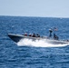 A rigid-hull inflatable boat (RHIB) is underway at high speed on open blue ocean water. The vessel carries multiple personnel in military/tactical gear and displays a United States flag at the stern. The boat is creating a significant wake and spray, indicating fast movement.