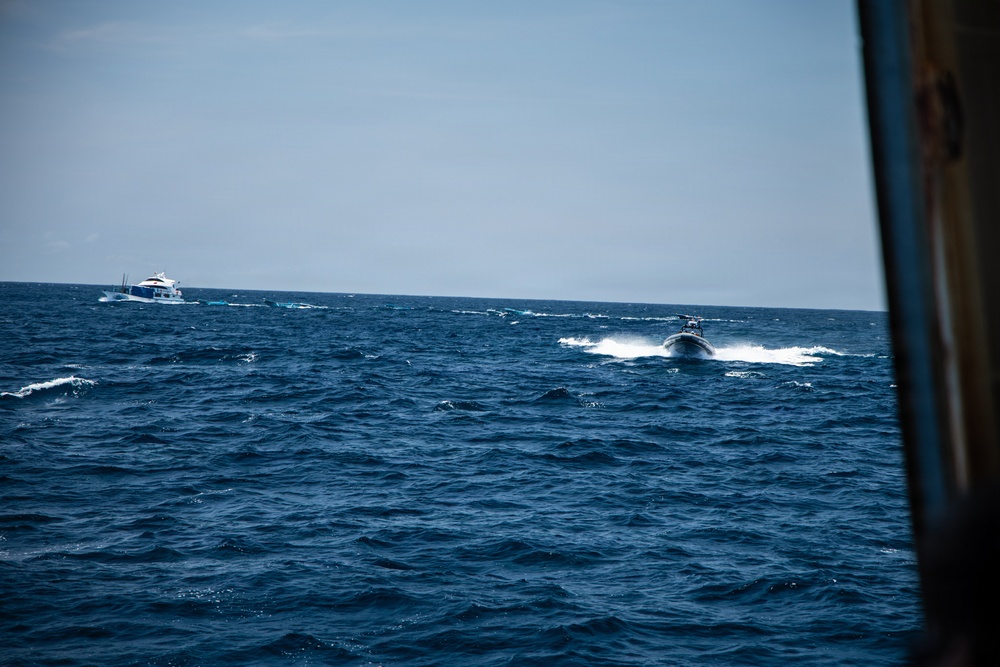 The image shows an open ocean scene with two vessels visible on choppy blue water. A smaller, fast-moving rigid inflatable boat (RIB) is in the foreground-middle distance, creating a white wake as it speeds toward a larger white motor yacht in the background. The photo appears to be taken from another vessel, with a partial view of a rusted hull or structure visible on the right edge.