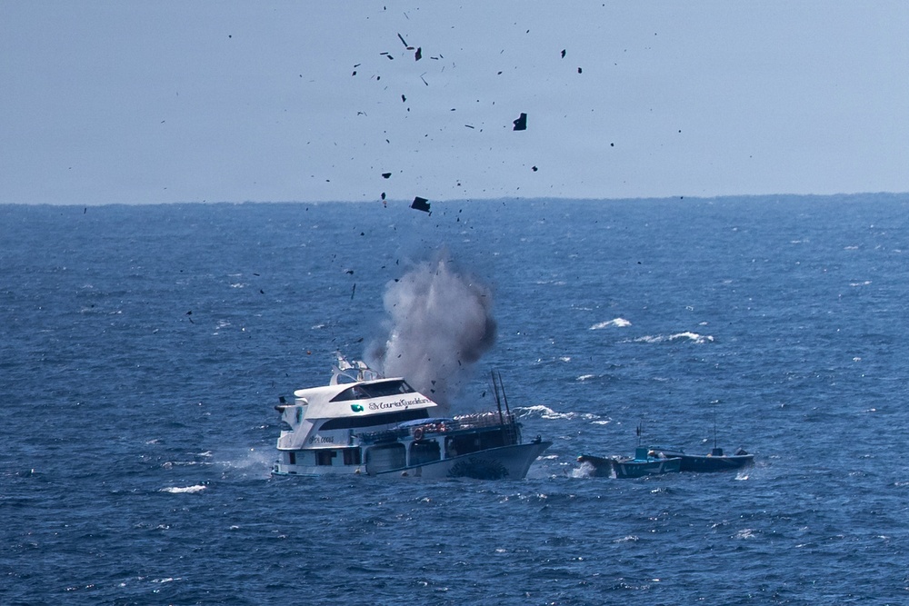 A white multi-deck vessel is experiencing an explosion or severe mechanical failure at sea, with debris and dark fragments being propelled into the air above the boat. A smaller vessel is visible nearby to the right of the main boat. The incident is occurring in open, choppy blue ocean water under a hazy sky.