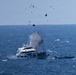A white multi-deck vessel is experiencing an explosion or severe mechanical failure at sea, with debris and dark fragments being propelled into the air above the boat. A smaller vessel is visible nearby to the right of the main boat. The incident is occurring in open, choppy blue ocean water under a hazy sky.