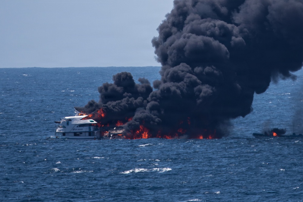 A large white multi-deck vessel is engulfed in intense flames on open water, producing a massive column of thick black smoke rising high into the sky. A smaller vessel is also visible to the right, appearing to also be on fire or heavily affected by the incident. The fire appears to be a serious maritime emergency occurring in calm, open sea conditions.