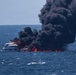 A large white multi-deck vessel is engulfed in intense flames on open water, producing a massive column of thick black smoke rising high into the sky. A smaller vessel is also visible to the right, appearing to also be on fire or heavily affected by the incident. The fire appears to be a serious maritime emergency occurring in calm, open sea conditions.
