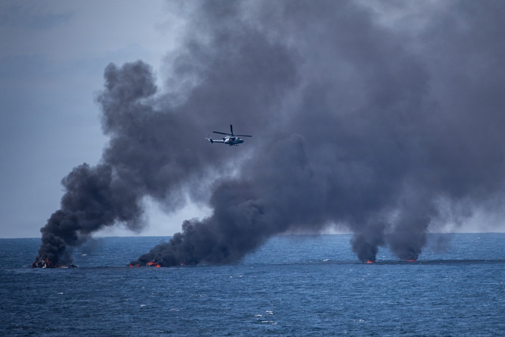 A military or coast guard helicopter flies over an open ocean scene where multiple fires are burning on the water surface, producing large columns of dark black smoke. The fires appear to be concentrated in at least two distinct clusters on the water. This appears consistent with a controlled burn operation or destruction of seized contraband at sea.