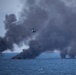 A military or coast guard helicopter flies over an open ocean scene where multiple fires are burning on the water surface, producing large columns of dark black smoke. The fires appear to be concentrated in at least two distinct clusters on the water. This appears consistent with a controlled burn operation or destruction of seized contraband at sea.