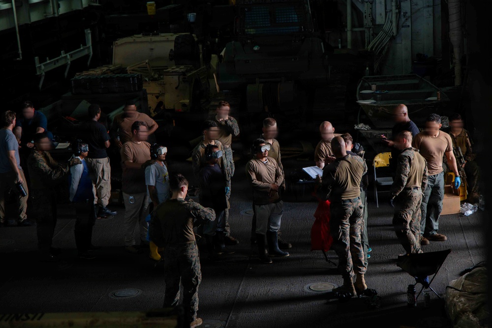 A group of military personnel, appearing to be U.S. Marines based on their camouflage uniforms, are gathered in what appears to be the well deck or vehicle bay of a naval vessel. Several individuals are wearing hearing protection and goggles, suggesting a pre-operation briefing or equipment check. Military vehicles are visible in the background of the dimly lit space.