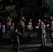 A group of military personnel, appearing to be U.S. Marines based on their camouflage uniforms, are gathered in what appears to be the well deck or vehicle bay of a naval vessel. Several individuals are wearing hearing protection and goggles, suggesting a pre-operation briefing or equipment check. Military vehicles are visible in the background of the dimly lit space.