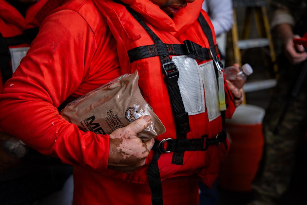 A person wearing a bright orange immersion suit and life vest is holding an MRE (Meal Ready-to-Eat) military ration packet. The individual's hand appears to have dirt or dried blood on it, suggesting they may have been involved in a rescue or emergency situation. The setting appears to be an indoor or sheltered area with other people visible in the background.