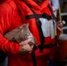 A person wearing a bright orange immersion suit and life vest is holding an MRE (Meal Ready-to-Eat) military ration packet. The individual's hand appears to have dirt or dried blood on it, suggesting they may have been involved in a rescue or emergency situation. The setting appears to be an indoor or sheltered area with other people visible in the background.