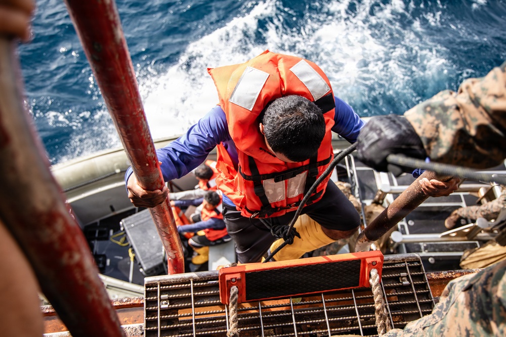 A person wearing an orange life vest and blue long-sleeve shirt is climbing a metal ladder or gangway from a smaller vessel onto a larger ship at sea. Several other individuals in orange life vests are visible in the boat below. A person in military camouflage uniform is visible on the right side, suggesting a possible maritime rescue or boarding operation.