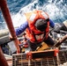 A person wearing an orange life vest and blue long-sleeve shirt is climbing a metal ladder or gangway from a smaller vessel onto a larger ship at sea. Several other individuals in orange life vests are visible in the boat below. A person in military camouflage uniform is visible on the right side, suggesting a possible maritime rescue or boarding operation.
