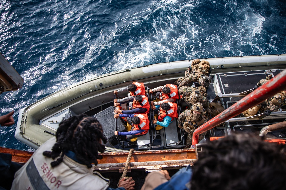 An aerial view of a maritime rescue or boarding operation showing individuals wearing orange life vests being assisted onto or transferred via a rigid inflatable boat (RIB). Military or law enforcement personnel in tactical gear are present on the right side of the vessel. The scene appears to take place in open, choppy ocean waters.