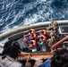 An aerial view of a maritime rescue or boarding operation showing individuals wearing orange life vests being assisted onto or transferred via a rigid inflatable boat (RIB). Military or law enforcement personnel in tactical gear are present on the right side of the vessel. The scene appears to take place in open, choppy ocean waters.