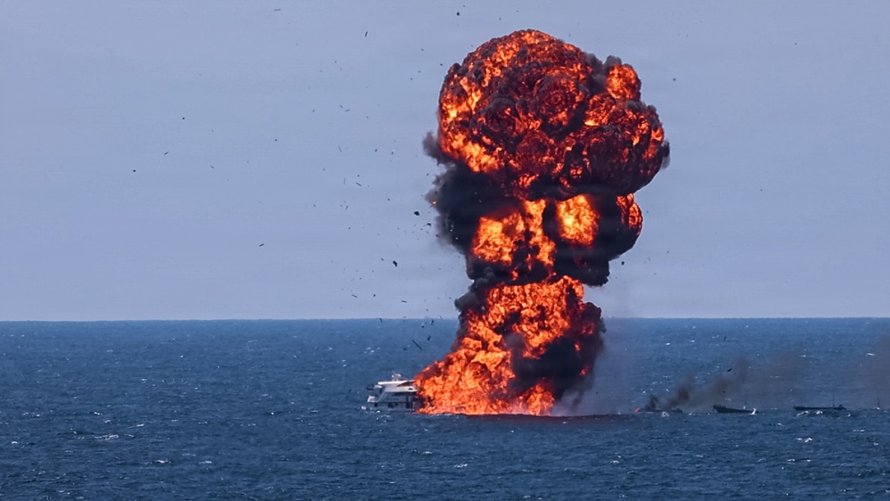 A large vessel on open water is engulfed in a massive explosion, producing a towering mushroom-shaped fireball with dense black and orange smoke. The explosion appears to originate from the deck or hull of a white boat, with the fireball rising high above the waterline. Several smaller vessels are visible nearby on the water.