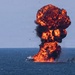 A large vessel on open water is engulfed in a massive explosion, producing a towering mushroom-shaped fireball with dense black and orange smoke. The explosion appears to originate from the deck or hull of a white boat, with the fireball rising high above the waterline. Several smaller vessels are visible nearby on the water.