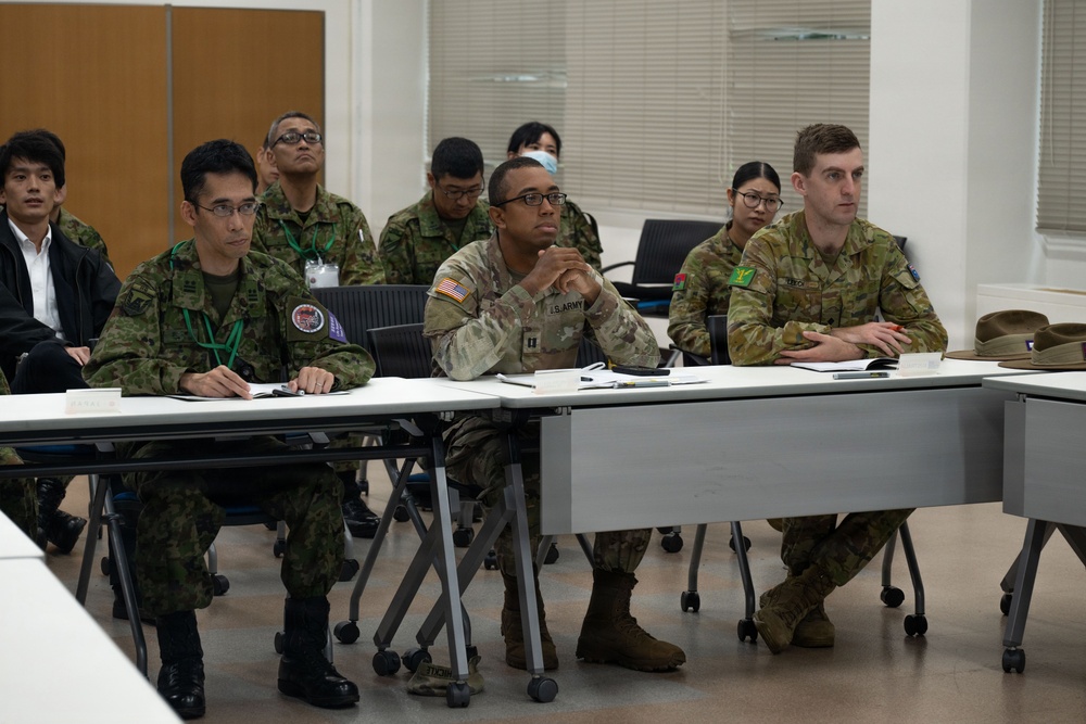 A multinational military briefing or training session is taking place in a classroom-style setting. Personnel from at least three different nations — identifiable by their distinct uniforms — are seated at tables and appear to be attentively listening to a presentation. The setting suggests a joint military exercise or cooperative training event.