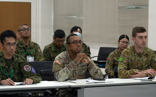 A multinational military briefing or training session is taking place in a classroom-style setting. Personnel from at least three different nations — identifiable by their distinct uniforms — are seated at tables and appear to be attentively listening to a presentation. The setting suggests a joint military exercise or cooperative training event.