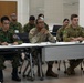 A multinational military briefing or training session is taking place in a classroom-style setting. Personnel from at least three different nations — identifiable by their distinct uniforms — are seated at tables and appear to be attentively listening to a presentation. The setting suggests a joint military exercise or cooperative training event.