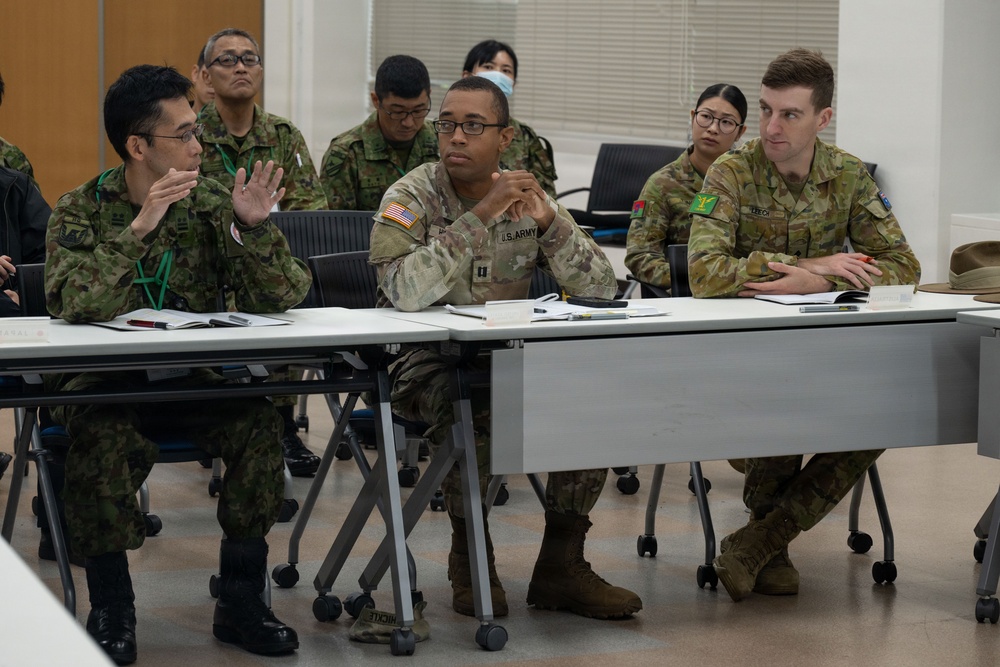 Military personnel from multiple nations are seated at tables in what appears to be a briefing or conference room setting. Visible uniforms include Japanese Ground Self-Defense Force (JGSDF), U.S. Army, and Australian Army personnel. The group appears to be engaged in a multinational military meeting or training exercise discussion.