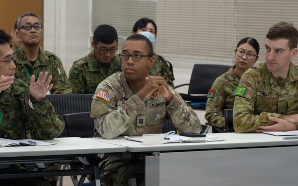 Military personnel from multiple nations are seated at tables in what appears to be a briefing or conference room setting. Visible uniforms include Japanese Ground Self-Defense Force (JGSDF), U.S. Army, and Australian Army personnel. The group appears to be engaged in a multinational military meeting or training exercise discussion.