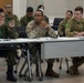 Military personnel from multiple nations are seated at tables in what appears to be a briefing or conference room setting. Visible uniforms include Japanese Ground Self-Defense Force (JGSDF), U.S. Army, and Australian Army personnel. The group appears to be engaged in a multinational military meeting or training exercise discussion.