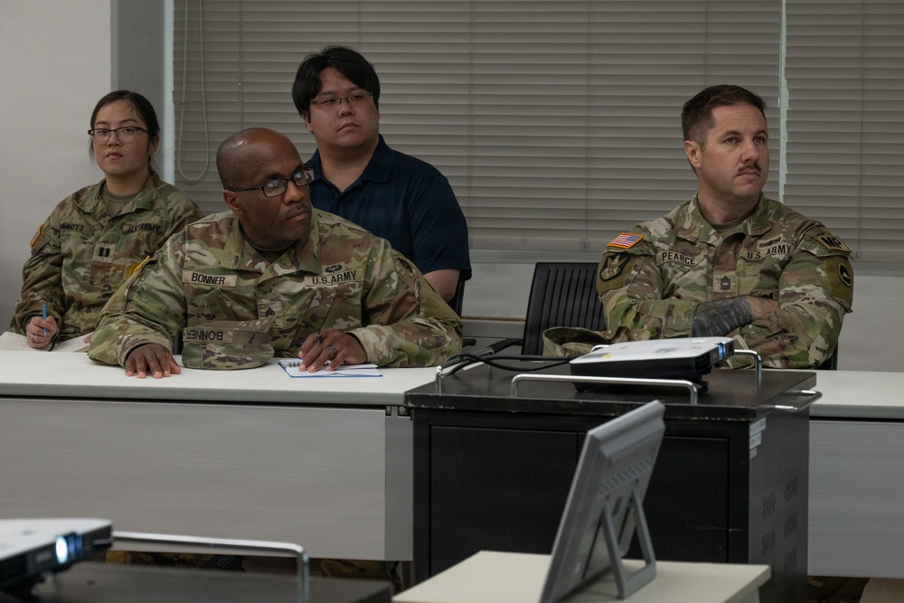 Three U.S. Army soldiers in OCP (Operational Camouflage Pattern) uniforms are seated at a table in what appears to be a classroom or briefing room setting. A civilian male in a navy polo shirt is seated behind them. The group appears to be attending a briefing, training session, or meeting.