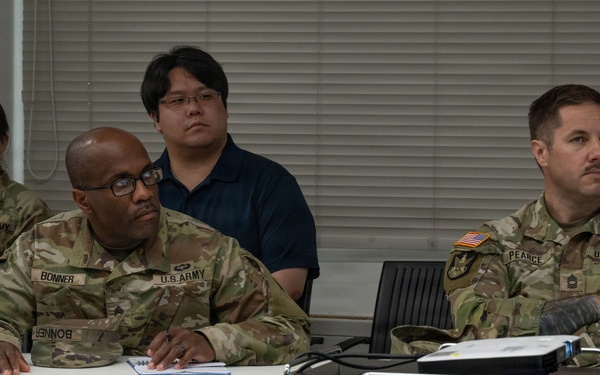 Three U.S. Army soldiers in OCP (Operational Camouflage Pattern) uniforms are seated at a table in what appears to be a classroom or briefing room setting. A civilian male in a navy polo shirt is seated behind them. The group appears to be attending a briefing, training session, or meeting.