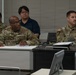 Three U.S. Army soldiers in OCP (Operational Camouflage Pattern) uniforms are seated at a table in what appears to be a classroom or briefing room setting. A civilian male in a navy polo shirt is seated behind them. The group appears to be attending a briefing, training session, or meeting.
