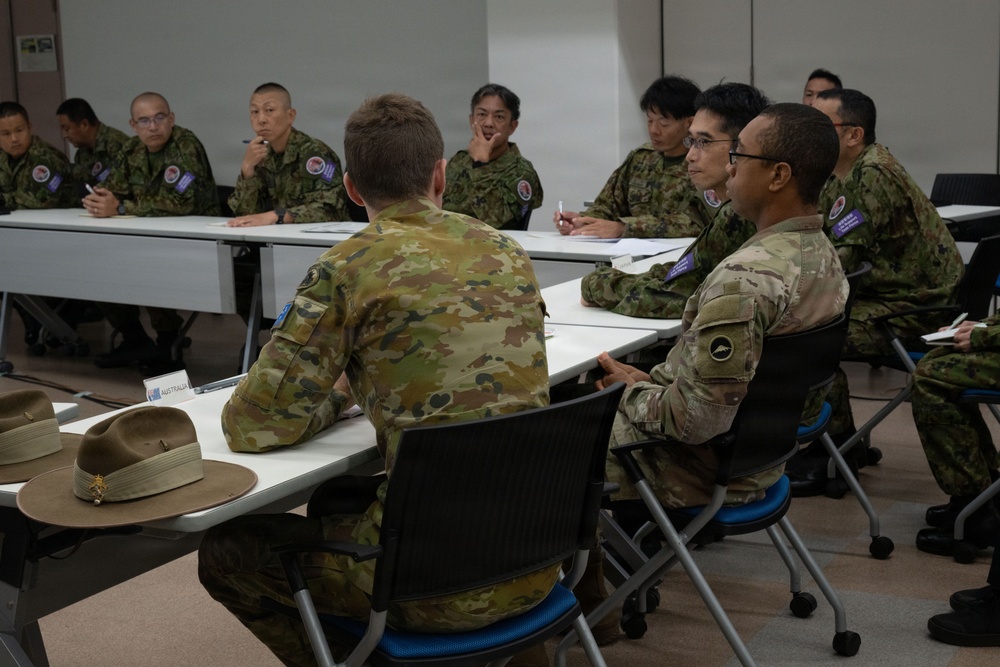 A multinational military meeting or briefing is taking place in a conference room setting. Personnel wearing different camouflage uniforms, including Australian Defence Force (ADF) and what appears to be Japan Self-Defense Force (JSDF) uniforms, are seated around tables facing each other. A name placard reading 'AUSTRALIA' is visible on the near table, along with a distinctive Australian slouch hat.