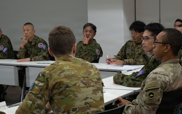 A multinational military meeting or briefing is taking place in a conference room setting. Personnel wearing different camouflage uniforms, including Australian Defence Force (ADF) and what appears to be Japan Self-Defense Force (JSDF) uniforms, are seated around tables facing each other. A name placard reading 'AUSTRALIA' is visible on the near table, along with a distinctive Australian slouch hat.