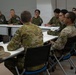 A multinational military meeting or briefing is taking place in a conference room setting. Personnel wearing different camouflage uniforms, including Australian Defence Force (ADF) and what appears to be Japan Self-Defense Force (JSDF) uniforms, are seated around tables facing each other. A name placard reading 'AUSTRALIA' is visible on the near table, along with a distinctive Australian slouch hat.