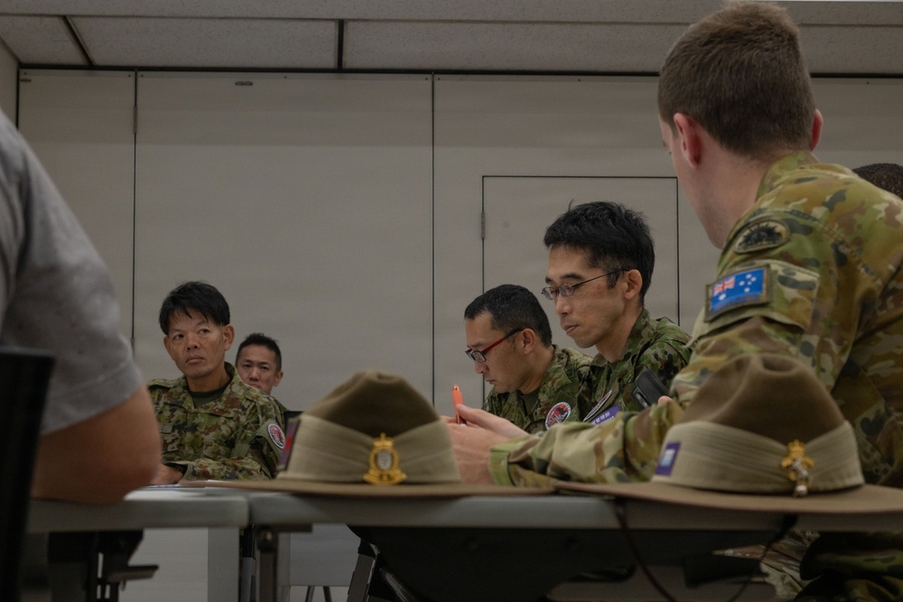 A military meeting or briefing session involving personnel from at least two nations — Australia and Japan — seated around a table in an indoor facility. The Australian soldier is identifiable by the distinctive Southern Cross flag patch on his multicam uniform, while the other personnel wear Japanese Ground Self-Defense Force camouflage uniforms. Slouch hats with badge insignia are placed on the table, indicating an Australian Army presence.